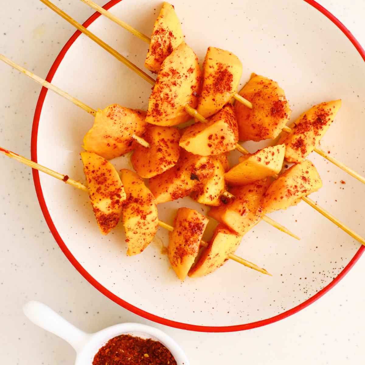 four individual wooden skewers containing mango tanghulu pieces on a white plate.