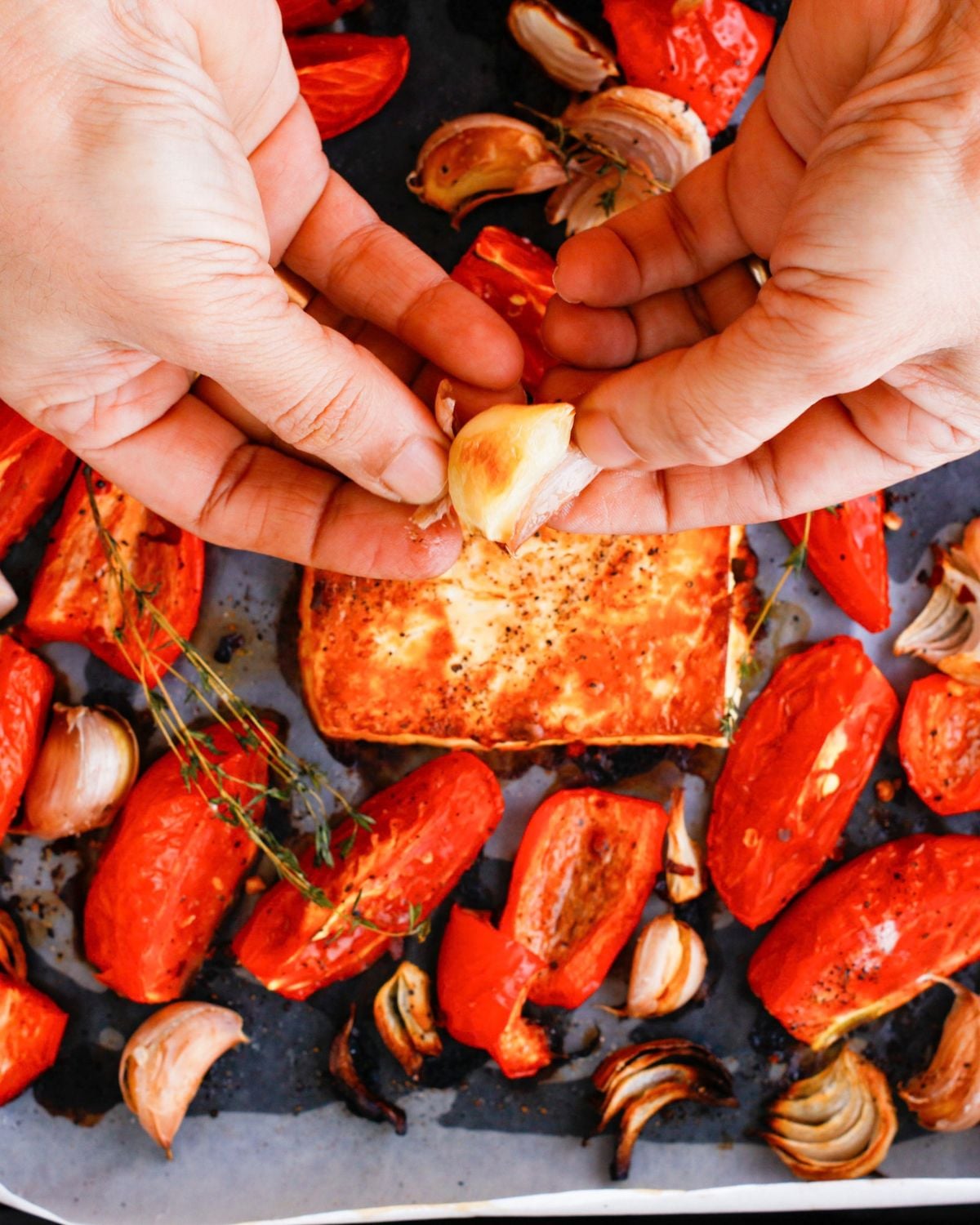 Peeling roasted garlic beside roasted vegetables and feta